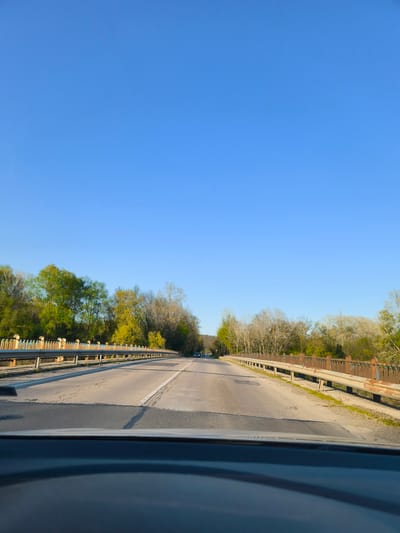 A forward-facing view from inside a car traveling on a bridge near Staro Oriahovo, Bulgaria. The road is asphalt and has a white line dividing the lanes. The bridge has old, weathered metal railings on both sides. Trees line both sides of the road, showing early spring foliage. The sky is clear blue. A car is visible in the distance ahead. The dashboard of the car taking the photo is visible in the foreground.