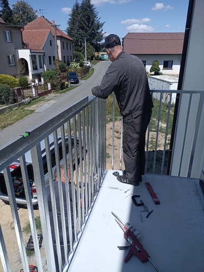 In Rataje, Czechia, on a sunny day, a man in dark work overalls and a baseball cap is seen on a balcony, facing away from the camera and towards a white railing. He appears to be working on the railing, with a level placed on top of it. Below him, another man in a grey shirt is partially visible, also working on the balcony. The balcony has a white floor and is surrounded by a metal railing. Tools and materials, including a caulk gun, a clamp, screws, and a level, are scattered on the balcony floor. In the background, a street with houses and trees lines the scene. A dark blue car is parked on the street. The overall impression is a construction or renovation project in progress.