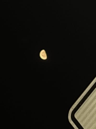 This image depicts an outdoor nighttime scene in Chuchuligovo, Bulgaria. The upper-left quadrant of the frame is dominated by a distinct, yellowish-white crescent moon, illuminated on its right side and appearing slightly diffused against the dark, black sky. Faint topographical textures are visible on the moon's illuminated surface. In the bottom-right corner, a partial view of a man-made structure is present, featuring a rounded corner. This object exhibits a light-beige or off-white surface with parallel dark lines running along its edge, creating a striped, textured appearance. No people, specific actions, or direct interactions are visible. The environment is characterized by the profound darkness of the night sky, with the moon serving as the primary visible celestial object.