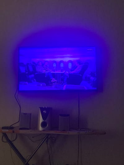 In the city of Lobnya, Russia, a flat-screen television is mounted on a wall, displaying a scene of passengers seated in an airplane cabin. The room is dimly lit, with a purple ambient light emanating from behind the television, casting a glow on the wall. Below the television, an ironing board serves as a makeshift shelf, holding two speakers and a small black device with a blinking light. Cables are visible, connecting the devices and trailing down the wall. The overall impression is one of a home entertainment setup in a sparsely furnished room.