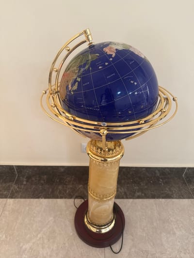 The scene depicts a large, ornate, floor-standing decorative globe situated indoors in the city of Aragats, Armenia. The globe features a deep blue sphere representing oceans, with continents and landmasses depicted in various natural stone colors, possibly inlaid (e.g., light brown, green). Gold-colored grid lines indicating latitude and longitude, along with place names such as "PACIFIC," "NORTH," "OCEAN," "Micronesia," and "Polynesia," are inscribed on the sphere.

The globe is cradled within an elaborate, highly polished gold-plated brass armillary sphere structure, consisting of multiple concentric rings and a central axis, some rings accented with small, clear, faceted gems. This assembly rests upon an ornate golden collar, which surmounts a cylindrical pedestal. The pedestal column appears to be crafted from a translucent, veined, creamy-yellowish stone, consistent with onyx. The column is adorned with additional decorative gold-colored bands.

The entire apparatus stands on a circular, polished dark wood base, resembling mahogany, which has a gold-colored metal ring at its junction with the stone column. A black electrical cable is visible on the floor, extending from behind the base.

The floor beneath the globe is tiled in two distinct sections: a lighter beige-grey marble-patterned tile in the foreground and a darker grey-brown marble-patterned tile behind it, separated by a horizontal grout line. The background consists of a plain, light beige or off-white wall. A standard white duplex electrical outlet is mounted on the wall behind the pedestal, slightly to the left of its vertical axis.

No individuals are directly visible in the scene, but a reflection, likely of the photographer and their device, is discernible on the glossy blue surface of the globe. No specific actions or interactions are depicted beyond the implied act of photography.