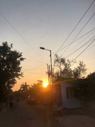 This is a photograph taken at sunset on a street in Sentul, Indonesia. The sky is a gradient of light blue and orange, with the sun near the horizon, casting a warm glow and lens flares. Overhead, a complex network of electrical wires crisscrosses the sky, supported by utility poles.
On the left side of the street, a large tree with dense foliage is silhouetted against the bright sky. Further down the street, there are several motorcycles, one of which is being ridden by a person. A dark-colored SUV is parked on the right side of the road.
Adjacent to the road on the right is a small building with a light-colored exterior and a green roof. Trees and other vegetation are visible throughout the scene, adding to the natural environment of the location. The road itself appears to be unpaved or in a rural setting.