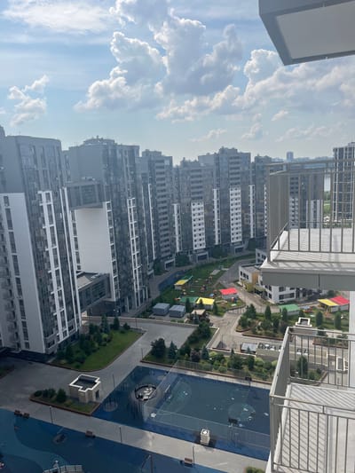 This is a high-angle, medium shot of a modern residential complex in Vysokaya Gora, Russia. The frame is dominated by tall, gray apartment buildings with many windows. In the foreground, a balcony railing is visible. Below, a courtyard features a sports court with blue surfacing and basketball hoops, a playground with small, colorful structures, and areas of green grass and trees. The sky above is partly cloudy. There are no people or animals visible in the image. The overall impression is one of a well-maintained, newly developed urban residential area.
