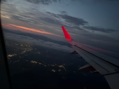 The image depicts a view from an airplane window at dusk. The scene is dominated by a dramatic sky with streaks of orange and pink from the setting sun, contrasting with dark, heavy clouds. Below, a sprawling city lights up the landscape, suggesting an urban or suburban area. The lights twinkle in organized patterns, hinting at roads and buildings. In the foreground, the wing of the airplane is prominent, painted red on its wingtip and bearing the text "Wizzair.com." The wing casts a shadow over the illuminated city. The overall atmosphere is one of travel and the transition from day to night, with a sense of vastness and elevation. The weather appears to be partly cloudy, with a clear view of the sunset. The location is identified as Vena di Maida, Italy, a detail that places the scene geographically. There are no people visible in the image. The image captures the beauty of a flight at twilight, highlighting the contrast between the natural beauty of the sky and the man-made lights of the city below, framed by the modern technology of the airplane.