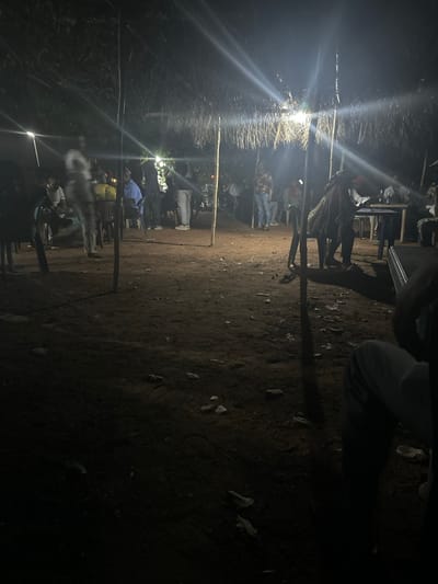 A dimly lit outdoor gathering at night. People are positioned within a rough-hewn enclosure marked by upright poles. A bright light source casts strong shadows on the ground. The ground is uneven and scattered with debris. Some individuals are seated on chairs, while others stand. The scene suggests a social event in a rural or less developed area.