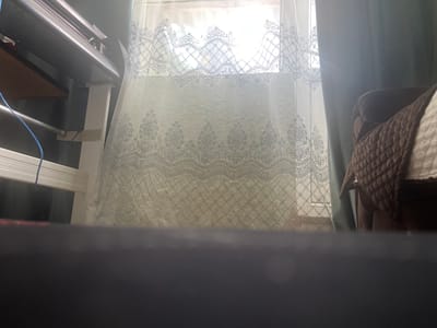 The image captures an indoor scene, likely a room, with a large window covered by decorative lace curtains. The sunlight streams through the window, creating a bright, diffused light. To the left, part of a large electronic device, possibly a printer or plotter, is visible, mounted on a white stand. A blue wire is attached to this equipment. To the right of the window, a dark teal curtain is partially drawn, and a portion of a brown quilted cushion or blanket is visible on a piece of furniture. The foreground is dominated by a dark, out-of-focus surface, which could be a table or floor. The scene is devoid of any people or visible activity. The location is identified as Novoselskoye, Russia.