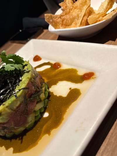 The image presents a close-up, elevated perspective of two food dishes arranged on a dark wooden table, presumably within a restaurant setting located in Sunrise, United States of America.

In the central-foreground, a white, rectangular ceramic plate showcases a stacked appetizer. This dish comprises visible layers, with a top layer of bright green, diced avocado, followed by a middle layer of reddish-pink, diced raw fish (consistent with tuna), and a bottom layer of small, green, spherical components, possibly edamame. The stack is garnished with dark, shredded nori seaweed and a sprig of fresh green cilantro on its apex. A viscous, dark green-yellowish sauce is artistically drizzled across the white plate surface around the food stack. Three distinct, small droplets of a reddish-orange condiment are strategically placed on the sauce and the plate.

In the upper-right background, a smaller, white, oval-shaped bowl is filled with a quantity of golden-brown, crispy, irregularly shaped items, consistent with fried wonton strips or thin chips.

The underlying surface is a dark brown wooden table. In the far-left background, a dark, indistinct silhouette of an individual is partially visible, wearing dark clothing. Adjacent to this figure, a dark, rectangular object with two small, white, circular reflections, consistent with a smartphone, rests on the table. The ambient lighting appears dim, characteristic of an indoor dining establishment.