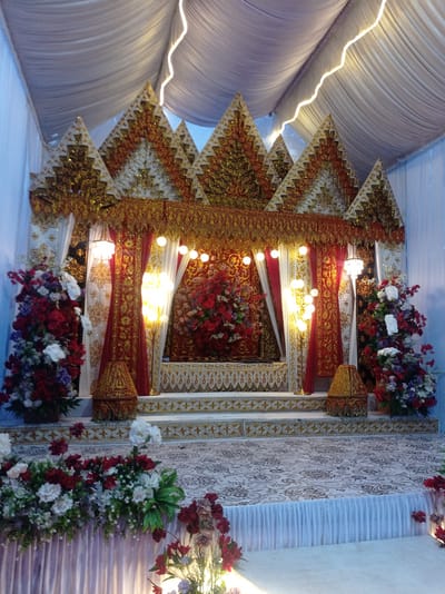 The image shows a stage decorated for a wedding ceremony. The stage is set up inside a tent with white fabric draped from the ceiling. The backdrop of the stage is adorned with intricate gold and red patterns, resembling traditional Indonesian architecture. There are also red curtains and white fabric panels. Several lamps are strategically placed to illuminate the stage, with a tall floor lamp on the right side. Large floral arrangements in red, white, and purple hues are positioned on either side of the stage and at the front. The stage floor is covered with a patterned carpet. The overall ambiance is festive and elaborate, indicative of a traditional wedding celebration. The description of the location as "Peudari, Indonesia" is assumed to be context provided for the image.