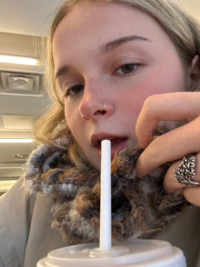 A young woman with fair skin and freckles poses with a white straw in her mouth. She wears a nose ring and a chunky silver ring. Her eye color is grey-green. A fuzzy, brown and grey scarf encircles her neck. The photo is taken at an angle, with the background showing a wall and doorway. A light fixture is visible in the upper left corner.