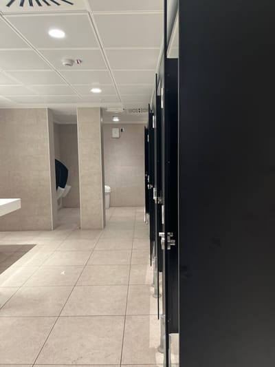 This image depicts a modern, empty public restroom in Dorno, Italy. The perspective is slightly angled, looking towards a row of restroom stalls. The scene is characterized by clean lines and neutral tones.
The main subjects are the restroom stalls, which feature dark, possibly black, partitions with chrome-like hardware for hinges and latches. At least three stalls are visible, with two toilets identifiable in the background. One toilet is within an open stall, and another is partially visible behind a low partition. In the foreground of one stall, a black object, possibly a bag or piece of clothing, lies on the floor.
The left side of the room features white paneled walls with integrated circular lights, illuminating the area. A red fire alarm or smoke detector is mounted on one of these panels, and a ventilation grille is visible further to the left. The floor throughout the restroom is covered in light-colored, square tiles, which also extend partway up the walls in the stall area. On a tiled wall in the background, a white dispenser (possibly for soap or paper towels) is mounted above a small white waste bin. The ceiling appears to be dark, contrasting with the lighter walls and floor.
No people are present, and no specific activity is taking place, suggesting the restroom is currently unoccupied. The lighting is artificial, and there is no indication of the time of day or outdoor weather. No visible text is discernible in the image. The overall impression is one of a clean, well-maintained, and functional public facility.