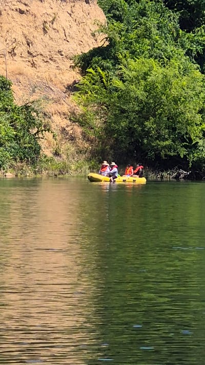 In the city of Carmichael, California, United States, a yellow raft is being paddled down a body of water. The raft carries four individuals, each wearing a life vest, two in white hats and shirts, and two in red and black life vests. The raft is positioned in the middle of the water, with a tree-covered embankment and a sandy cliff face visible in the background. The water reflects the surrounding foliage and the sky, creating a dappled, green and yellow surface. The sun appears to be shining, casting shadows and highlighting the textures of the natural landscape. The scene suggests a leisurely outdoor activity, likely on a river or lake.