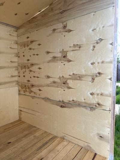 The image captures the interior of a basic, unfinished wooden structure, likely a shed, large container, or raised garden bed, viewed from above and slightly to the side.
The primary subject is the wooden paneling that forms the walls and floor of this structure. The prominent central and left wall is constructed from light-colored plywood, highly visible for its numerous dark brown knots. From many of these knots, darker, vertical streaking patterns descend, indicative of sap or moisture stains. Small fasteners, such as nails or screws, are visible securing this plywood panel. A thin, dark line, possibly a string or wire, runs vertically across this panel.
To the right, a different wall is visible, composed of horizontally aligned, rough-hewn wooden planks. These planks exhibit varied tones and textures, suggesting rustic or repurposed wood, possibly from pallets. The floor of the structure is also made of light-colored plywood, with visible fasteners.
The structure appears to be open at the top, revealing a dark-colored rim along its upper edge. Beyond this, a small section of green vegetation, possibly grass or bushes, is visible against a bright, light-colored background, suggesting an outdoor setting during daytime.
No people or activities are present in the image. There is no visible text. The construction materials suggest a functional, utilitarian purpose. The image provides no specific visual cues to its purported location in Kuda, Russia.