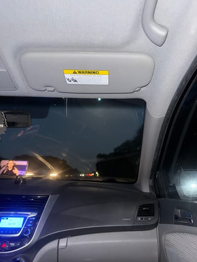 The image shows the interior of a car at night. The view is from the passenger seat, looking forward. The dashboard, center console, and part of the driver's side door are visible. A smartphone is mounted on the dashboard, displaying a video of a person. The windshield reflects the dark sky and the headlights of other vehicles on the road. The car appears to be moving. A warning sticker is visible on the passenger side visor. The car is located in Manheim Township, United States of America.