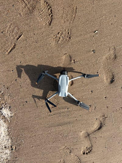 A grey and white drone rests on a sandy beach in Ak Dobo, Kyrgyzstan. The drone is in the center of the frame, lying on its side with its four propellers extended. Its body is rectangular with rounded edges, and it has a black camera lens at the front. The drone appears to be a DJI Mavic 3, identifiable by the branding on its body.

The sand is a light brown color, with a scattering of small pebbles and dried plant matter visible along the top edge of the image. There are also several footprints pressed into the sand, suggesting that people have been walking on the beach. The footprints are of varying sizes and shapes, indicating the presence of both adult and possibly child footprints. Some of the prints are clear, while others are partially obscured by sand.

The scene is illuminated by bright sunlight, casting a long shadow from the drone and its propellers. The shadow suggests that the sun is relatively low in the sky, indicating either early morning or late afternoon. The weather appears to be clear and sunny, with no clouds visible.

The overall impression is one of a peaceful, outdoor setting. The drone's placement on the sand suggests it may have just landed or is about to take off. The presence of footprints implies recent human activity, but the drone itself is stationary. The image captures a moment of stillness on a natural landscape.