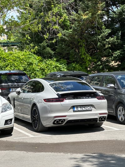 The image shows a parking lot in the city of Варна, Bulgaria, featuring several parked vehicles. The most prominent vehicle is a white Porsche with a Bulgarian license plate number "B 9292 CA." Other cars are parked adjacent to the Porsche, including a dark SUV and a silver crossover. The background consists of dense green trees and foliage. The parking lot appears to be located in a residential or commercial area. The scene is captured during daylight hours with clear sunlight. The focus is primarily on the rear of the Porsche and the surrounding parked cars.