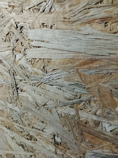 The image is a close-up, high-angle shot of Oriented Strand Board (OSB). The surface is composed of compressed wood chips and strands, varying in color from light beige to brown, with some darker areas indicating moisture or age. The texture is rough and uneven, with visible grain patterns and individual wood pieces. There are no people, animals, or discernible objects other than the OSB itself. The lighting is somewhat dim, highlighting the textured surface. The image does not contain any information that would allow for the localization to the city of Жалпақсай, Kazakhstan.