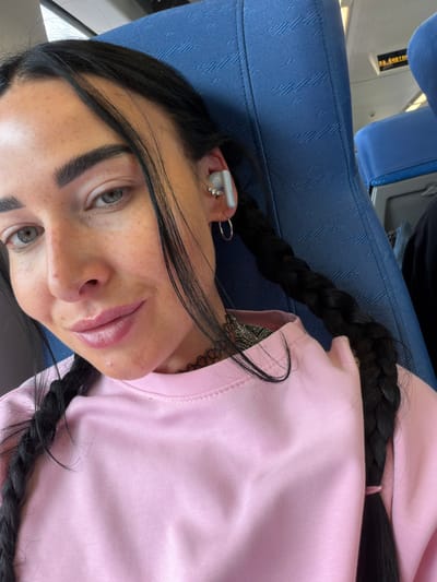 The media file is a close-up, selfie-style portrait of a young woman with dark hair styled in two braids, wearing a pink long-sleeved top. She is seated, and the background suggests an indoor setting, possibly public transportation given the blue upholstered seating visible. The lighting appears to be natural, indicating daytime. The woman has light-colored eyes, noticeable freckles across her face, and she is wearing small hoop earrings in her left ear. Her lips are glossy.
A small piece of white text is visible on what appears to be an earbud in her left ear, which reads "HUAWEI". There is also visible text on a yellow sign in the upper left corner of the image, in Russian: "ЗА БОРТОМ" (ZA BORTOM), meaning "Outside".
The overall mood is casual and relaxed, with the woman looking directly at the camera. The scene is likely within Russia, given the provided location context.