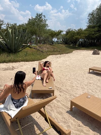 A woman in a bikini is lying on a lounge chair on a sandy beach in Pueblo Nuevo de Jasso, Mexico. Another woman stands near the lounge chair and holds a smartphone up to take a picture of her. There are other lounge chairs and a side table nearby. Trees and agave plants line the edge of the beach in the background. The sky is partly cloudy.