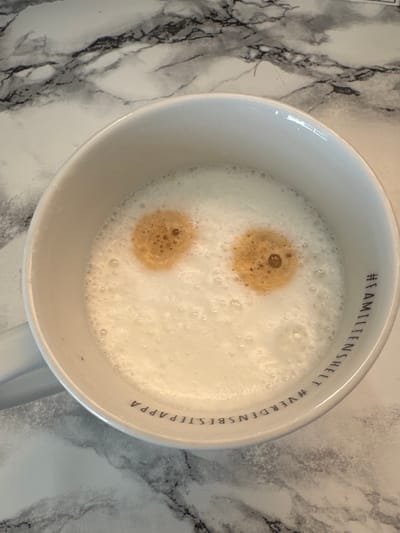 An overhead, medium shot shows a white mug filled with a frothy, light-colored beverage, likely a latte or cappuccino, resting on a white marble-patterned surface. Two circular dollops of darker liquid, resembling coffee or espresso, are positioned in the foam, forming what appears to be eyes. The mug has text printed in black around its rim. The text reads "#FAMILIESHELT #VERDENSBESTEPAPPA". The scene is indoors, possibly in a kitchen or dining area, in Moelv, Norway. No people are visible in the image.