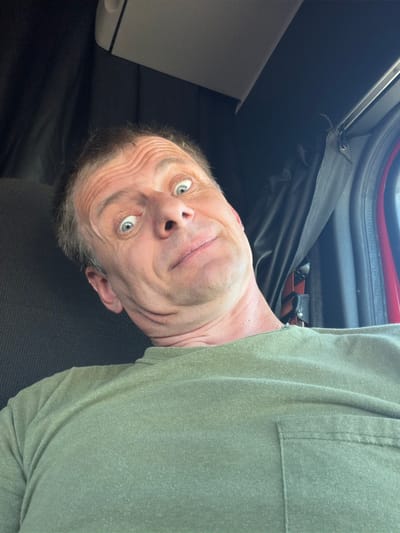 A man is taking a selfie inside a truck cab. He is wearing a green t-shirt and has his head tilted, making a surprised expression with wide eyes and raised eyebrows. The background shows the interior of a truck, including dark curtains and a red accent on the right side. The image appears to be taken during the day, with natural light coming in. The man's face is the central focus of the image.