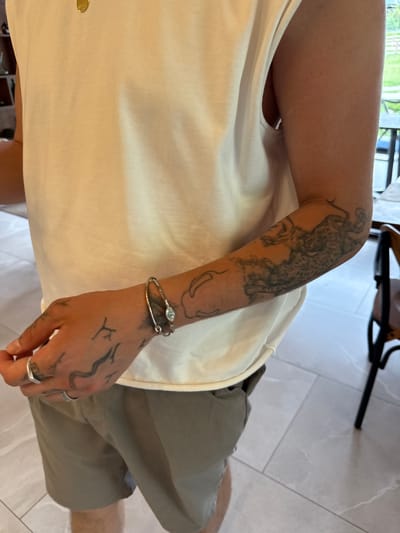 A close-up shot shows a person from the chest down, wearing a light-colored sleeveless t-shirt and khaki shorts. The person has several tattoos on their left arm and hand, and is wearing a silver bracelet on their left wrist and two silver rings on their left index finger. The background includes a tiled floor and a glimpse of outdoor scenery with trees and a white fence, suggesting a location in Dsegh, Armenia.