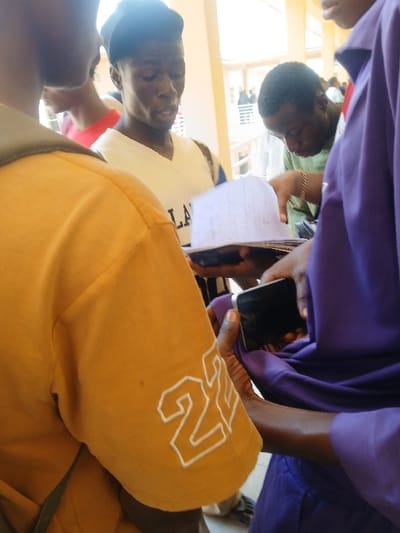 The scene, reportedly in Ugi Majowel, Nigeria, depicts a group of at least five individuals gathered in an indoor or covered outdoor setting. In the immediate foreground, the back of a person wearing a mustard yellow short-sleeved shirt with "22" outlined in white on the right sleeve is partially visible. Central to the image is a young man with dark skin, wearing a white sleeveless top with visible dark lettering (partially reading "LA") and a dark cap, looking downwards with his mouth slightly open. To his right, a person in a purple shirt holds a black smartphone horizontally in their right hand, while their left hand is positioned near a stack of white papers or a booklet. Further to the right and slightly behind, another young man in a green shirt, wearing a gold-colored bracelet on his right wrist, leans forward, also observing the papers. Multiple indistinct figures are present in the background, along with architectural elements such as light-colored pillars and railings. The primary action involves multiple individuals collectively viewing and interacting with shared documents.