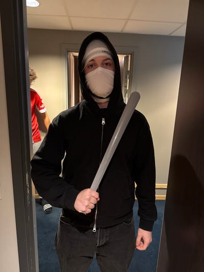 An indoor, portrait-oriented photograph captures a person standing within a doorway, looking directly at the camera. The individual wears a black hooded sweatshirt with the hood up, dark pants, and what appear to be Adidas slides on their visible left foot. A white face mask covers the lower half of their face, and a white headband with "Calvin Klein" text is visible across their forehead, just above their eyes. The person holds a light-colored, possibly plastic, bat diagonally across their body.

In the background, out of focus, another person wearing a red shirt is partially visible through a further doorway. The setting features light-colored walls and a blue carpeted floor, indicative of an indoor space like a hallway or room. The lighting appears to be artificial. The overall composition suggests a posed shot, possibly for a costume or dramatic effect. The location is Jessheim, Norway.
