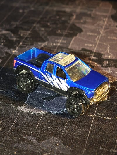 A blue toy pickup truck is photographed on a dark surface that appears to be a map, possibly of the world, with faint geographical details visible. The truck has a detailed design with silver graphics on the side and a clear plastic light bar on the roof. It has black, off-road style wheels and fender flares. The lighting in the image creates highlights and shadows, suggesting a close-up shot with focused lighting. No people or actions are depicted. The image does not contain any information that can be used to identify the location as Dörpen, Germany.
