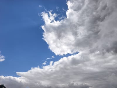 The image shows a sky with both blue and cloudy areas. A significant portion of the sky is a bright blue, while a large cluster of white and grey clouds occupies the right side of the frame. The clouds appear dense, with varying shades of grey suggesting depth and possibly rain-bearing potential. A small portion of a green structure is visible in the lower left corner. The location is the Jezreel Valley Regional Council, Israel.