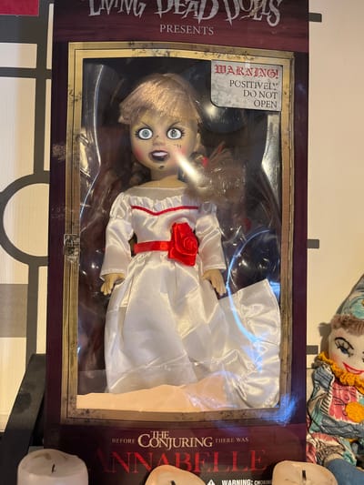 An Annabelle doll, dressed in a white satin gown with a red sash and rose, is displayed in its original packaging. The box, featuring the "Living Dead Dolls Presents" logo and "Before the Conjuring There Was Annabelle" text, has a warning label that reads "WARNING! POSITIVELY DO NOT OPEN." The doll's eyes are wide and blue, and it has dark eye makeup and red lips. The doll is positioned centrally in the frame, inside a clear plastic window. To the right of the Annabelle doll packaging is a colorful, handmade cloth doll with blue eyes and a red smile. In the foreground, the tops of two white candles are visible. The background consists of a patterned wall with vertical and horizontal lines. The scene appears to be indoors, possibly in a home or retail setting. There is no indication of the city of Schwäbisch Hall, Germany.