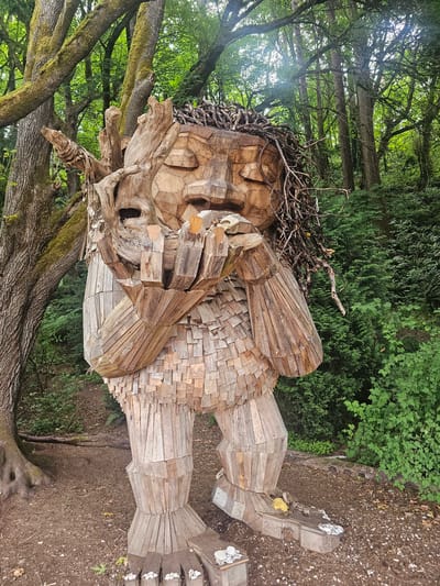 The image depicts a large wooden sculpture of a troll in a forest setting. The troll is made of many wooden planks and pieces, giving it a textured appearance. Its arms are raised and holding a piece of wood or stone to its face, and its eyes are closed. The troll's hair appears to be made of twigs and branches. The sculpture is situated on a dirt path surrounded by lush green trees and foliage. In the foreground, at the troll's feet, are several small white stones. The overall scene is one of an outdoor art installation within a natural environment. This sculpture is located in the Ballard neighborhood of Seattle, Washington, United States, specifically in the Golden Gardens Park.