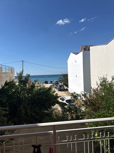 The image captures a view of Limenaria, Greece, on a clear, sunny day. The scene is framed by a white railing in the foreground, with greenery and trees in the mid-ground. Beyond the trees, a street lined with parked cars leads towards the sea. The ocean is calm and blue, with a small boat visible in the distance. Two white buildings with modern architecture dominate the right side of the frame, one of which has a red-tiled roof and a chimney. The sky is a vibrant blue with a few wispy clouds. The overall impression is one of a tranquil coastal town.