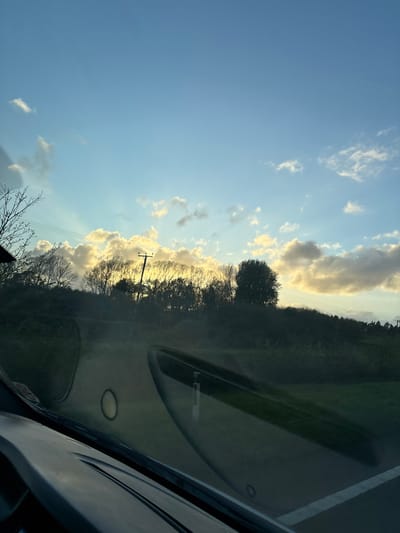 The image, taken from inside a vehicle, shows a view of the sky, trees, and a roadside landscape. The location is likely in Pentre, United Kingdom, given the prompt's instructions. The sky is blue with scattered clouds, some of which are illuminated by the sun. A line of trees and foliage runs along the horizon. A utility pole is visible among the trees. The foreground includes the interior of the vehicle, with the dashboard and windshield partially obscuring the view. There are no people visible in the image.