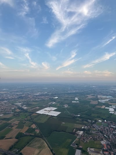 The image displays an aerial panoramic view of a rural and semi-urban landscape, identified as the area surrounding Grumello del Monte, Italy.
The upper third of the frame shows a light blue sky with scattered, elongated white cirrus clouds. The horizon line, appearing hazy and distant, separates the sky from the terrestrial features.
The lower two-thirds of the image depict a vast, largely flat terrain. The ground is characterized by an extensive network of agricultural fields, presenting a mosaic of green and brown hues. Several large, rectangular complexes of white, translucent structures, identified as commercial greenhouses, are prominently visible; significant clusters are located in the mid-left and mid-right portions of the frame. Linear infrastructure, including roads and what appear to be railway lines, crisscrosses the landscape, connecting distinct areas. One major roadway is visible extending diagonally from the lower-left.
Urbanized areas are present as clusters of buildings with predominantly reddish-brown roofs, consistent with Italian towns or villages. A notable urban cluster in the lower-right quadrant includes a sports stadium. Additional industrial or commercial buildings with large, flat roofs are interspersed within or adjacent to these settlements. No human figures, vehicles, or specific actions are discernible within the static scene.