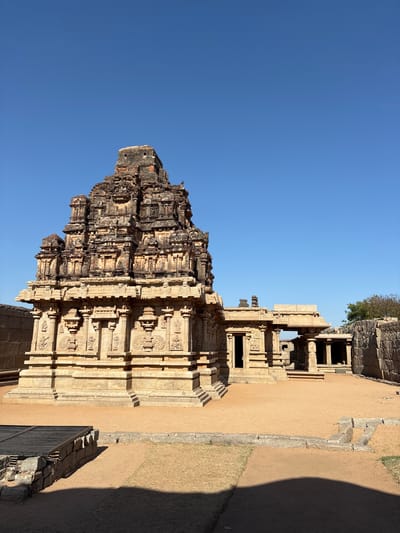 The image shows the Hazara Rama Temple in Kamalapura, India. The structure is a large, ornate stone building with multiple tiers and detailed carvings. The sky is clear and blue. The surrounding area appears to be dry and dusty, with other stone structures in the background.