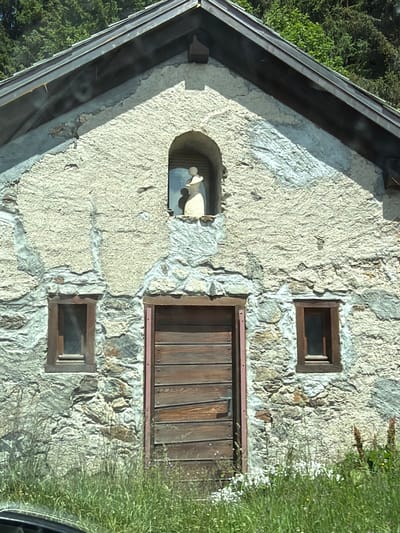 The image shows the front facade of a stone and stucco building, likely a small chapel or hermitage, set against a backdrop of green trees. The building has a pitched roof covered with dark material. A small, arched niche is centered on the upper part of the facade, containing a light-colored statue of a person wearing a hat. Below the niche, there is a large wooden door with horizontal planks. Two small windows with wooden frames are located on either side of the door, one on the left and one on the right. The walls are constructed of rough-hewn stones and a textured stucco finish. The foreground is overgrown with tall green grass and scattered wildflowers. The scene appears to be located in a rural or mountainous area, consistent with the setting of Vallorcine, France.