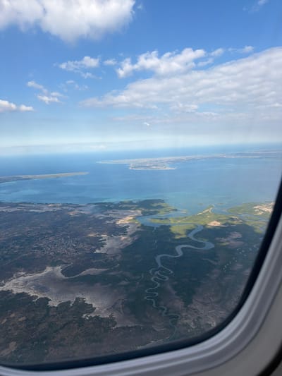 This media file captures an aerial view from an airplane window, showcasing a landscape characterized by a winding river, lush vegetation, and a distant body of water. The scene is bathed in the bright light of daytime, with a clear blue sky dotted with scattered white clouds, suggesting fair weather.

The dominant subjects are the natural elements of the earth and sky. A prominent river snakes its way through a dense green forest, creating intricate patterns against the terrain. Patches of lighter-colored land, possibly indicating drier areas or salt flats, break up the expanse of greenery. In the distance, a vast expanse of calm blue water, likely the ocean or a large bay, stretches towards the horizon. A narrow strip of land, possibly an island or a peninsula, is visible within the water, with a settlement or a cluster of buildings on its far side.

The image portrays a tranquil and awe-inspiring scene, emphasizing the grandeur of natural landscapes from a unique perspective. The time of day appears to be morning or early afternoon, judging by the bright illumination and the quality of the shadows, which are not prominently visible but implied by the contrast in the terrain. The location is identified as Madondo, Mozambique, which aligns with the presence of coastal features and potentially tropical vegetation. No text is visible in the media file.