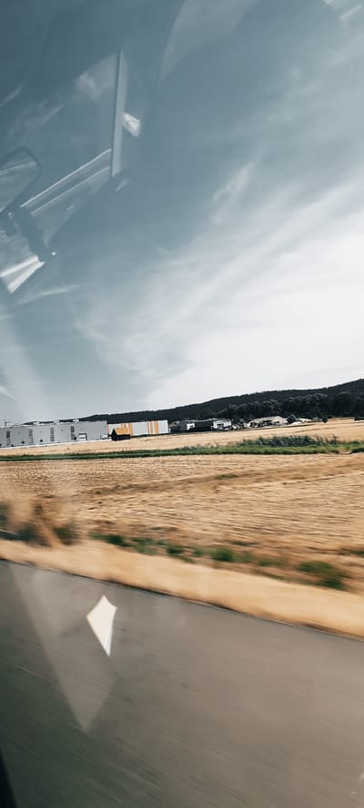 The image captures a blurred outdoor scene, likely taken from a moving vehicle. In the foreground, a grey, blurred surface resembling a road is visible. Above the road, a field of dry, golden grass is out of focus. Further in the background, a long, low-slung building complex with a white facade and orange accents is situated. To the right of the complex, a small, dark, gable-roofed structure stands in a field. Behind the buildings, a dark line of trees forms a hill. The sky above is a pale blue with wispy clouds. Reflections of the vehicle's interior, including a white structure and parts of the windshield, are visible in the upper portion of the image. The scene is likely in the vicinity of Winterbach, Germany, given the industrial architecture and rural landscape.