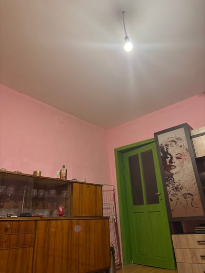 The image depicts an interior scene, possibly a room in a residential building in Kardam, Bulgaria. The walls are painted pink, and the ceiling is a light neutral color. A bare light bulb hangs from a cord in the ceiling, illuminating the space.
To the left, a wooden cabinet with glass doors displays an assortment of glassware, including wine glasses and tumblers. Below the glass-fronted section are wooden drawers and cabinets. On top of the cabinet, a bottle of what appears to be balsamic vinegar and other decorative items are visible.
To the right of the cabinet is a bright green door with glass panels in the upper section. Next to the door, leaning against the wall, is a framed picture of Marilyn Monroe in a black and white artistic rendering. Further to the right, a light-colored dresser with drawers is partially visible. A metal drying rack is positioned near the green door. The floor appears to be wooden. The overall impression is of a lived-in space with a mix of vintage and modern elements. There are no people or active interactions depicted in the image.