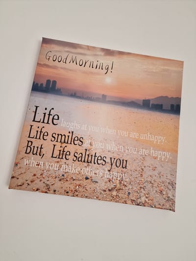 A canvas print displays a motivational quote against a sunset beach scene. The quote reads, "Good Morning! Life laughs at you when you are unhappy. Life smiles at you when you are happy. But, Life salutes you when you make others happy." The background of the print features a sandy beach with scattered pebbles and shells, a calm body of water, and a distant city skyline silhouetted against a vibrant orange and pink sky. The image is cropped to show the canvas print at an angle, with a plain white background surrounding it. No people are visible in the image. The location of Ovoshtnik, Bulgaria, is not depicted or referenced in the image.
