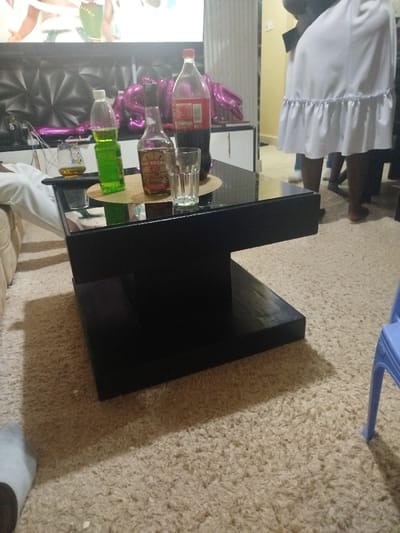 The image captures an indoor scene, likely a living room, characterized by a light-colored, shaggy carpet covering the floor. In the center of the frame is a modern, black coffee table with a reflective glass top.

On the coffee table, several beverages are arranged on a circular, woven placemat. These include:
*   A clear plastic bottle containing a bright green liquid, possibly a soft drink or mixer.
*   A brown glass bottle labeled "TOP SECRET" in white text, featuring an illustration of a figure with a hat, suggesting an alcoholic spirit like rum or whiskey.
*   A large plastic bottle of Coca-Cola with its distinctive red label and dark liquid.
*   An empty, clear drinking glass with a ribbed texture.
*   Another short glass containing a yellowish-brown liquid, likely an alcoholic beverage, perhaps neat or mixed.

To the left, a section of a light-colored sofa or armchair is visible, along with a person's arm wearing a white sleeve, partially obscured.

The background reveals a large screen, possibly a television, displaying blurry, colorful content, which could be an animated scene or cartoon. Below this screen, on a dark, patterned surface, several pink metallic balloons, possibly forming part of a celebratory message, are visible. This hints at a special occasion or party.

On the right side of the background, several people are partially seen. One person prominently wears a white, short-sleeved top or dress with a ruffled hem, standing with their bare feet visible. Other legs and feet, some bare and some in socks, are also discernible, indicating multiple individuals are gathered in the space. A blue plastic chair is partially visible on the far right.

Shadows from the coffee table are cast on the carpet, suggesting an indoor light source. The overall impression is that of a casual social gathering or party, possibly in the evening, with drinks being served.