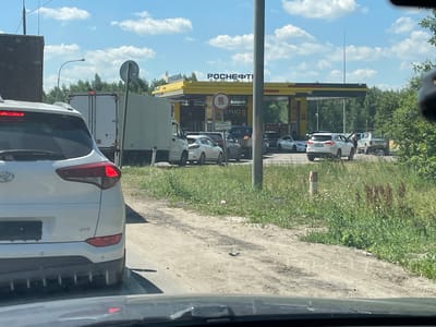 A white SUV is stopped in traffic on a dirt road, with the rear of the vehicle facing the camera. The SUV is a modern model, with its brake lights illuminated. Behind the SUV, a white box truck is also stopped. In the background, a yellow gas station with the "ROSNЕFT" logo is visible, suggesting the vehicles are likely waiting in line to refuel. Several other cars are also lined up at the gas station. The scene appears to be during the day, with a bright sky and scattered clouds, indicating fair weather. The surrounding area is covered in green grass and trees. A traffic sign with a red circle and a truck icon is posted near the gas station. The overall atmosphere seems to be one of a typical traffic jam at a gas station.
