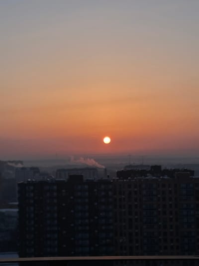 This image depicts an elevated panoramic view over the city of Elektrougli, Russia, at either sunrise or sunset. The sky transitions from a pale blue-gray at the top to vibrant orange and red hues towards the horizon. A prominent, bright, circular, orange-white sun is situated in the central sky area. The immediate foreground features the dark silhouettes of multiple multi-story residential buildings, some dark gray-black and others a muted brown-orange, obscuring the lower portions of the distant cityscape. A widespread atmospheric haze or fog blankets the mid-distance urban landscape. Within this hazy layer, distinct white plumes of smoke are observed rising from at least two separate, indistinct sources. No individuals, specific actions, or interactions are discernible. The environment is an urban setting characterized by densely packed residential structures and visible industrial or heating emissions.