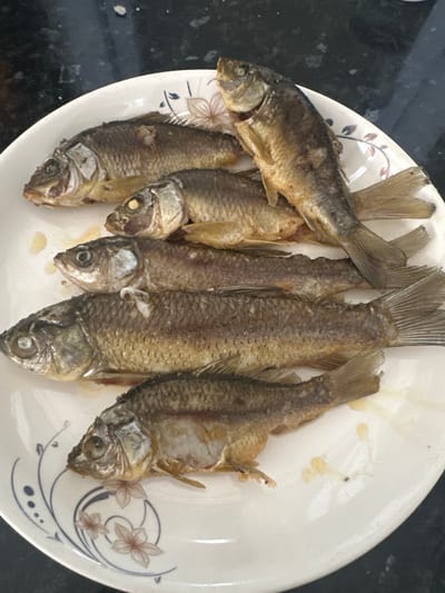 The image shows a plate of several small, whole, fried fish. The fish are arranged on a white plate with a decorative floral pattern. The plate itself appears to be white ceramic, with delicate lines and flower motifs in shades of brown and blue. The fish are golden brown, indicating they have been fried. Their eyes are visible, and their scales are distinct. Some of the fish have their fins spread, while others are more tightly packed. The background is dark and appears to be a countertop or table, with some subtle texture. There are a few small droplets of oil or sauce on the plate around the fish. The lighting suggests an indoor setting, possibly a kitchen or dining area. The overall impression is of a prepared meal, likely ready to be served. The crops provide closer views of the fish, their tails, heads, and the plate's design, highlighting the details of the food and the dish. There is no visible text in the image.
