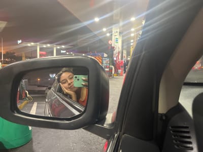 A young woman sits in a car at a Repsol gas station in Lurín, Peru, at night. She is visible in the driver's side mirror, holding a light green smartphone and resting her head on her hand, seemingly taking a selfie. The gas station, illuminated by bright overhead lights, features several gas pumps and attendants. The surrounding area appears urban, with other buildings and streetlights visible in the background.
