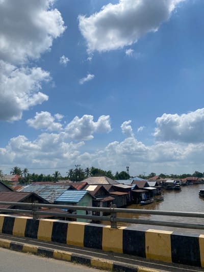 The image shows a view of the Berangas Timur village in Indonesia. The village is characterized by houses built on stilts over a brown river. The houses have a variety of roofing materials, including corrugated metal in shades of brown, red, and blue, as well as some with tiled roofs. Some houses have wooden walls and appear to be constructed with simple materials. Several small boats are visible on the river, some moored near the houses and others navigating the water.
In the foreground, a concrete barrier painted with alternating black and yellow stripes marks the edge of what appears to be a bridge or elevated walkway. Beyond the houses, a line of green trees and palm trees can be seen, with some taller structures, possibly utility poles or a water tower, interspersed among them. The sky is a clear blue, filled with numerous fluffy white cumulus clouds. The overall impression is of a tranquil, tropical village scene.