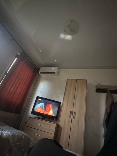 The image shows a dimly lit bedroom interior, angled from the top-left corner down to the bottom-right corner. A white ceiling with a central dome light fixture is visible in the top-left. The walls are light beige. Adjacent to the ceiling is a wooden rack with metal hooks. On the right, a light brown wooden dresser with metallic handles is situated, and a mounted flat-screen television is positioned to its left. Below the television, a portable air conditioner unit is placed on the floor near a window covered by a rust-colored curtain. A bed with a patterned comforter is partially visible in the bottom-right of the frame. The scene appears to be indoors. There is no explicit indication of this image's location in Pirdop, Bulgaria.