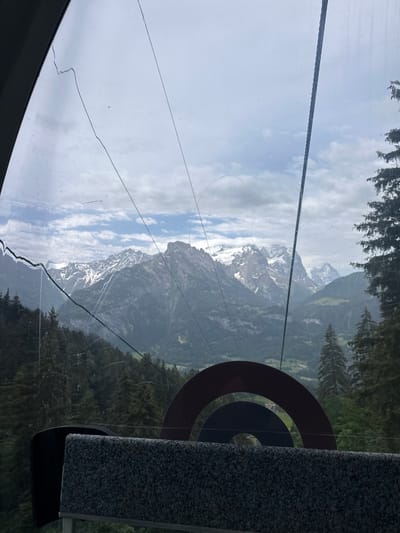 This image was taken from inside a cable car ascending towards Hasliberg Reuti, Switzerland. The view through the cable car's window reveals a majestic mountain landscape. Snow-capped peaks dominate the horizon, with rugged, dark grey mountains and green forested slopes below. The sky is overcast with scattered clouds. In the foreground, the interior of the cable car is visible, including a seat with a textured grey fabric and part of a dark grey frame. There are visible cracks and scratches on the cable car window, and a thick cable, likely part of the ski lift mechanism, runs diagonally across the frame.