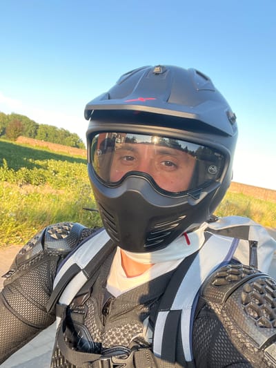 A person wearing a full-face helmet, protective armor, and a white shirt is visible from the chest up, facing the camera. The helmet is black with a clear visor and a black lower face guard. The armor covers the shoulders and chest. The background shows a sunlit grassy hill with trees and a brick wall, under a clear blue sky. The setting appears to be outdoors.