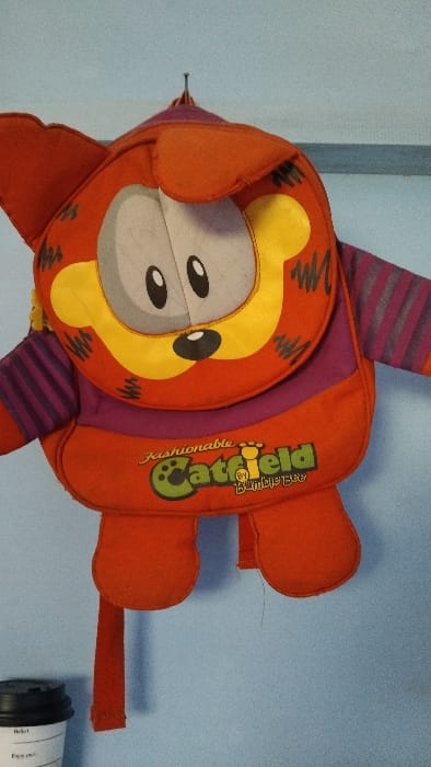 The image shows a red, orange, purple, and yellow backpack with a cat face on it. The backpack is hanging on a blue wall. The backpack has a cat face with two black eyes, a black nose, and a black mouth. The words "fashionable Catfield Bumibersama" are written on the backpack. The backpack is hanging from a hook on the wall. The image was taken in Kecamatan Panjalu, Indonesia, which is located at latitude: -7.143, longitude: 108.278. A black disposable coffee cup with the words "Hello, Enjoy your day" on it can be seen in the bottom left corner of the image. The backpack is slightly dusty. The image was likely taken indoors.